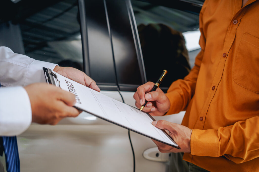 Asian clients engage with a car insurance agent beside a wrecked vehicle. They examine the policy details, assess the damage, and finalize paperwork related to the insurance claim and repairs.
