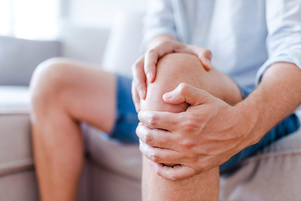Man suffering from knee pain sitting sofa. A mature man massaging his painful knee. Man suffering from knee pain at home, closeup. Pain knee