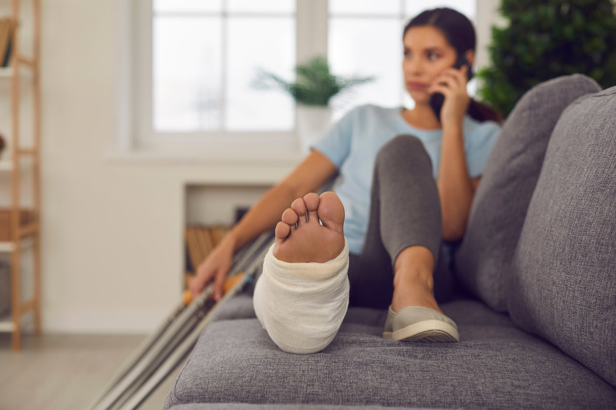 Young woman with broken leg in cast lying relaxing on sofa with crutches nearby and talking on phone at home, selective focus. Injury, trauma, recovery, rehabilitation concept