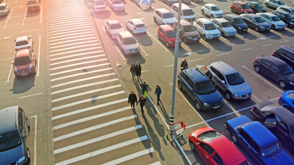 Pedestrian crossing and parking lot next to the entrance of a mall. Aerial. People go to the mall.