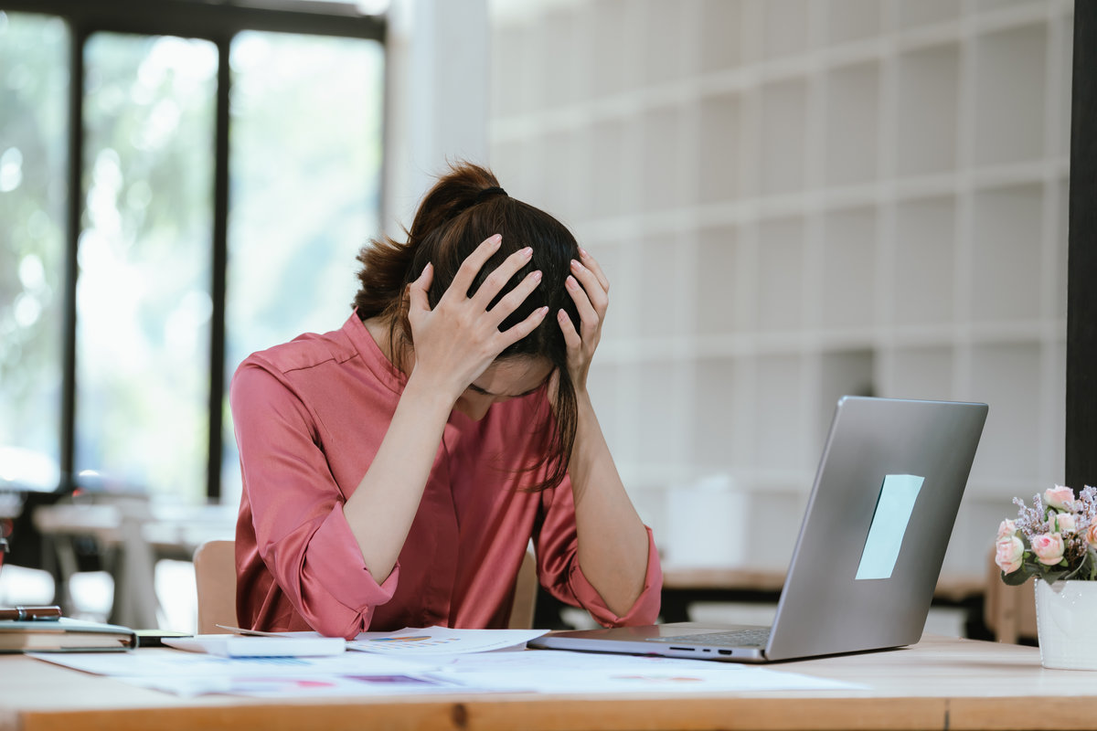 A stressed businesswoman at her desk holds a calculator and rubs her eyes while reviewing financial documents beside a laptop in a modern office.